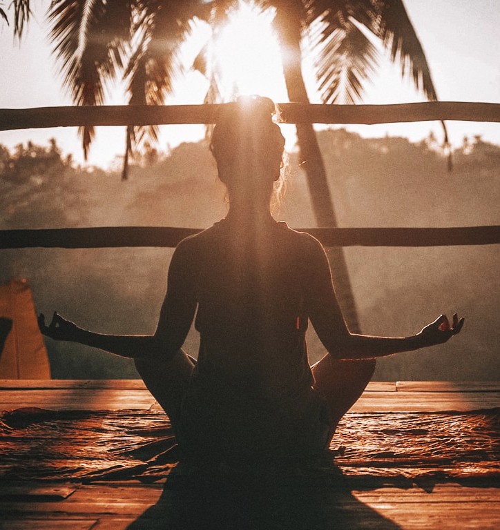Silhouette of a person meditating cross-legged on a wooden deck at golden sunrise with palm trees